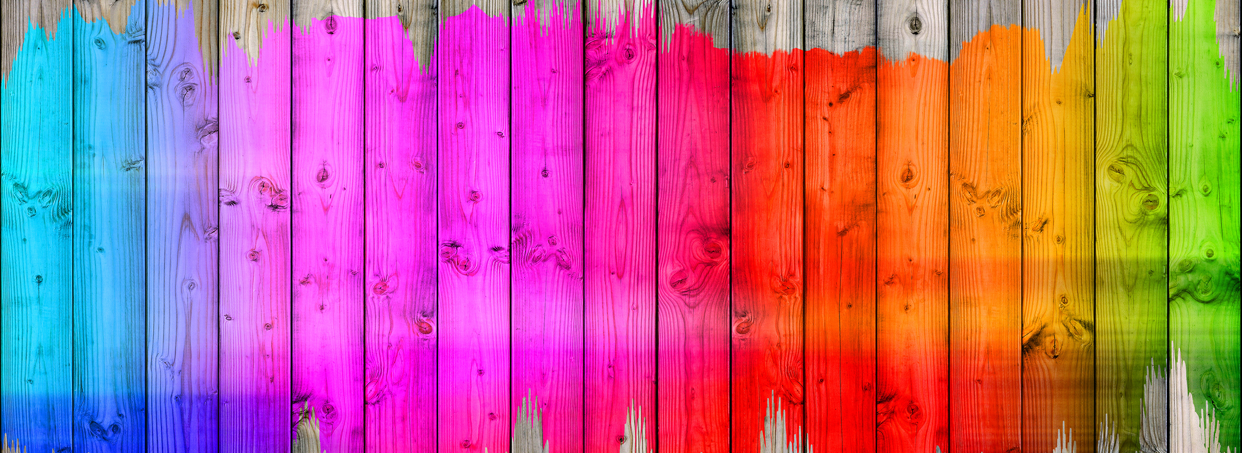 Das Bild zeigt eine hölzerne Wand oder einen Zaun, der in kräftigen Regenbogenfarben bemalt ist. Die vertikalen Holzlatten sind von links nach rechts in Blau, Violett, Pink, Rot, Orange, Gelb und Grün gestrichen. Symbolbild für Farbige Akzente Zuhause.