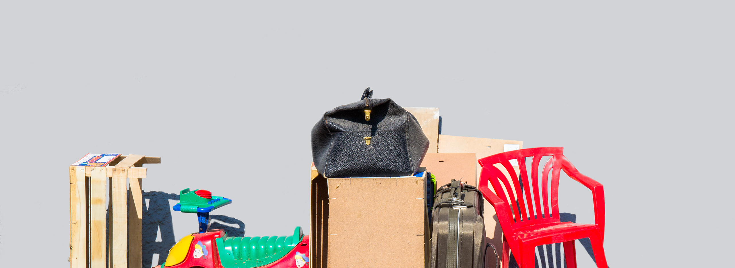 Ein Stapel ausgemusterter Gegenstände steht am Straßenrand: ein roter Plastikstuhl, eine schwarze Tasche, Holzkisten, ein Kinderfahrzeug und Kartons. Symbolbild für Entrümpeln oder Ausmisten.