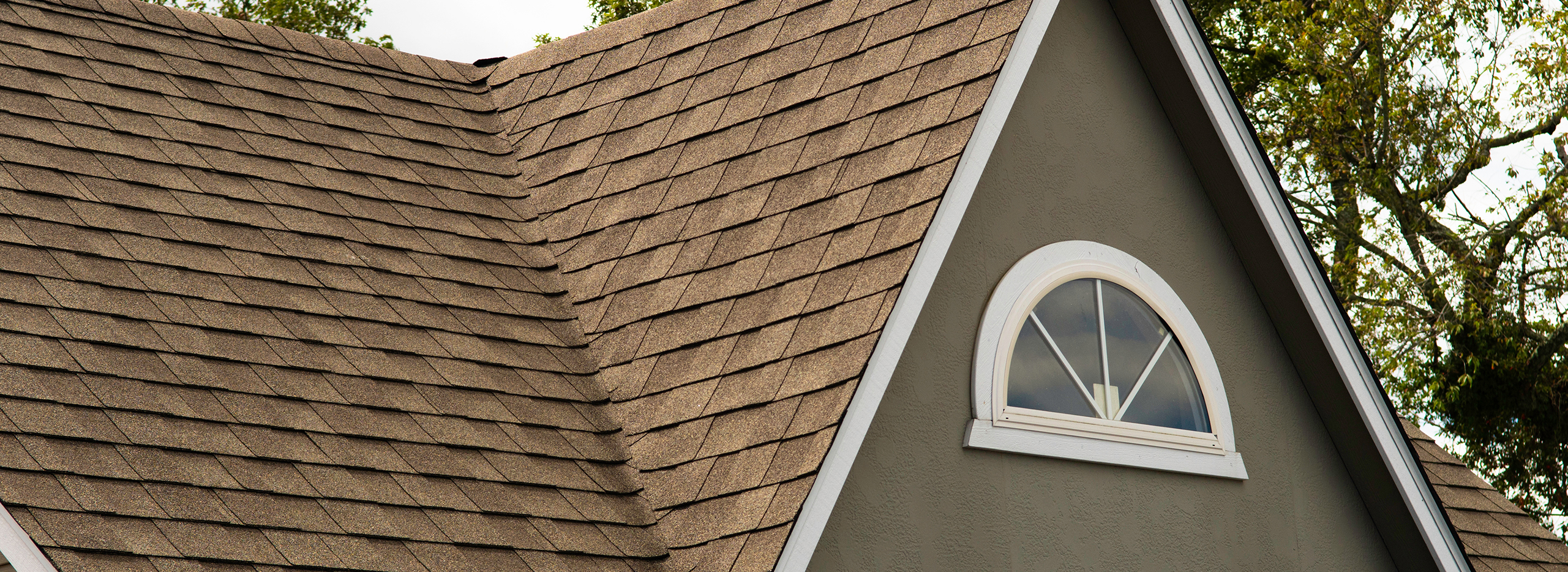 Dach eines Hauses mit braunen Schindeln und einem halbrunden Fenster im Giebel. Die Aufnahme zeigt die Dachstruktur und ist ein Symbolbild für verschiedene Dachformen.