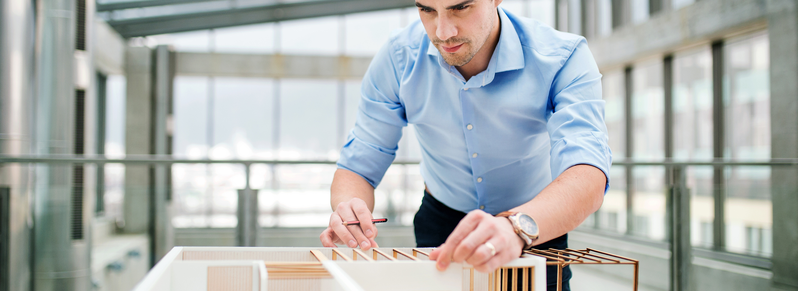 Ein Mann in einem hellblauen Hemd arbeitet konzentriert an einem Architekturmodell. Er hält einen Bleistift in der Hand und misst oder zeichnet Details nach. Symbolbild für Planung, Architektur oder Bauwesen.