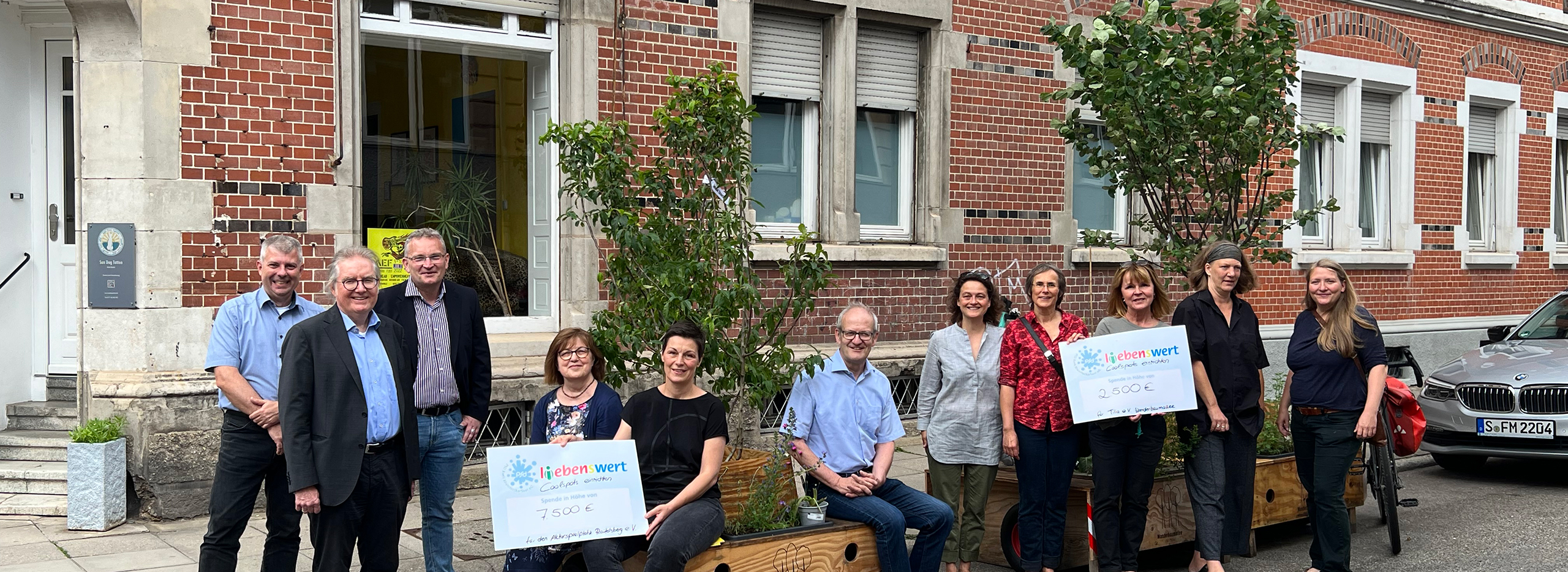 Gruppenfoto vor einem Backsteingebäude mit mehreren Personen, die zwei große Spendenschecks in den Händen halten, flankiert von bepflanzten Holzkübeln und kleinen Bäumen – symbolisch für eine Spendenübergabe der Stiftung PSD L(i)ebensWert.