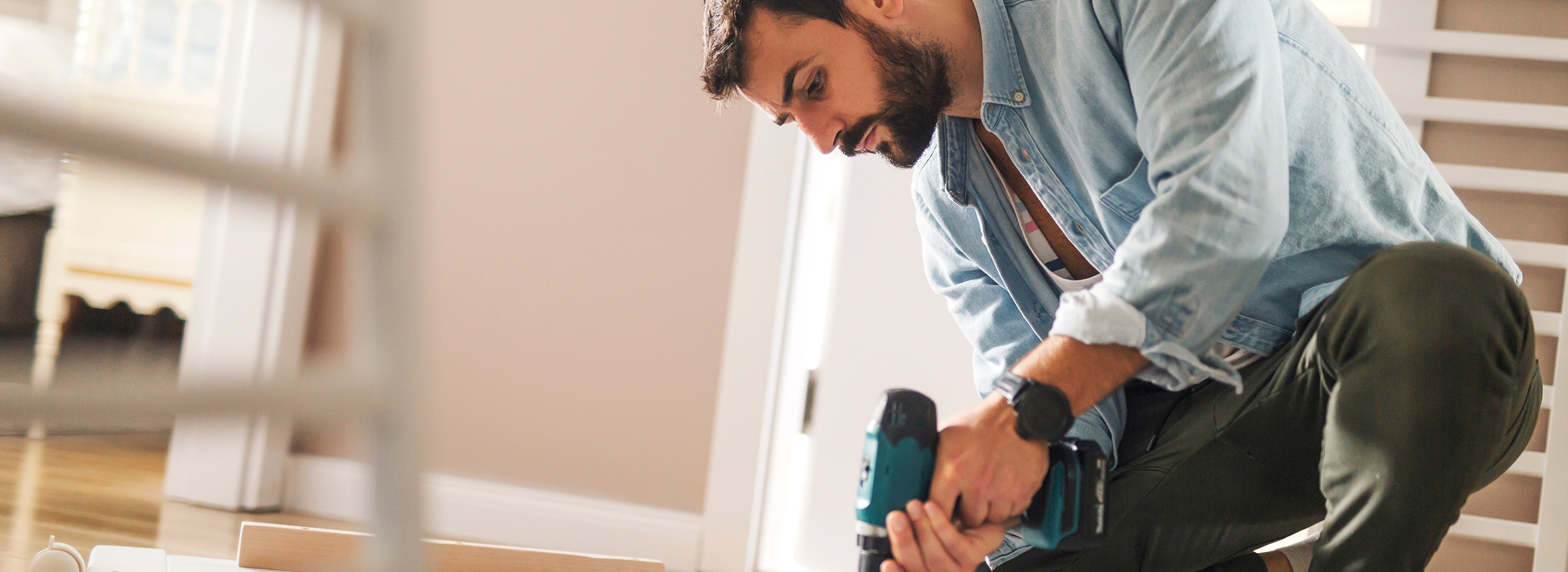 Ein Mann kniet auf dem Boden und arbeitet konzentriert mit einem Akkuschrauber an einem Möbelstück. Er trägt ein hellblaues Jeanshemd und olivgrüne Hose. Symbolbild für: Haften beim Heimwerken
