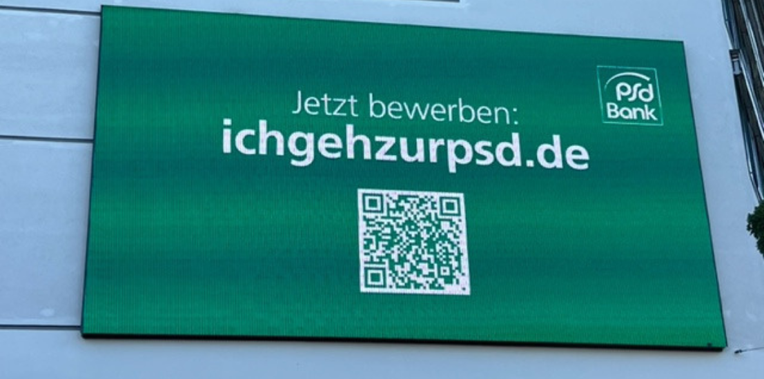 Eine große digitale Werbetafel an einer Hausfassade. Auf grünem Hintergrund steht in weißer Schrift: jetzt bewerben: ichgehzurpsd.de