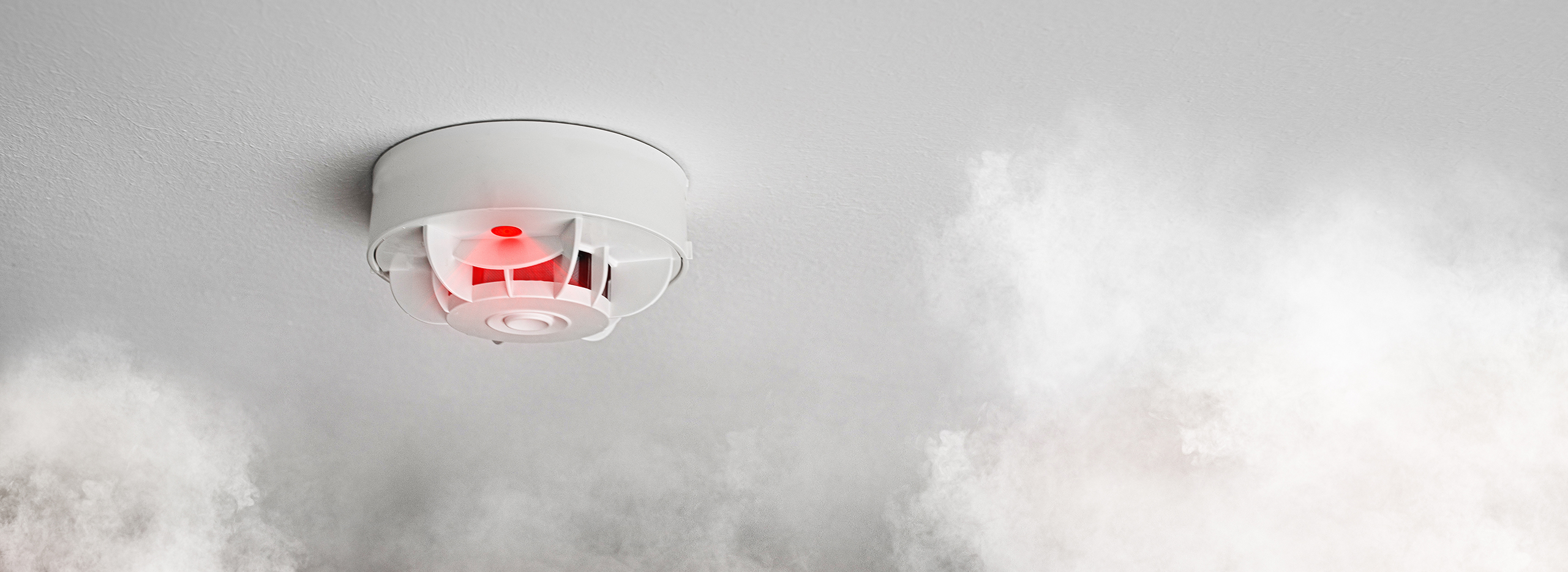 Das Bild zeigt einen Rauchmelder an der Decke, der aufgrund von aufsteigendem Rauch ausgelöst hat. Symbolbild für Wohnungsbrand.