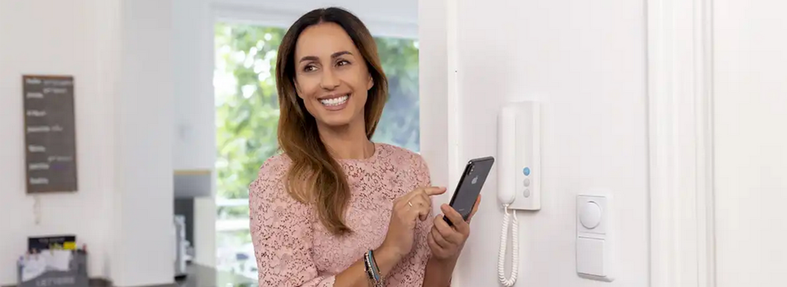 Das Bild zeigt eine Frau, die mit einem Smartphone neben einer modernen Gegensprechanlage im Hausflur steht und lächelt.