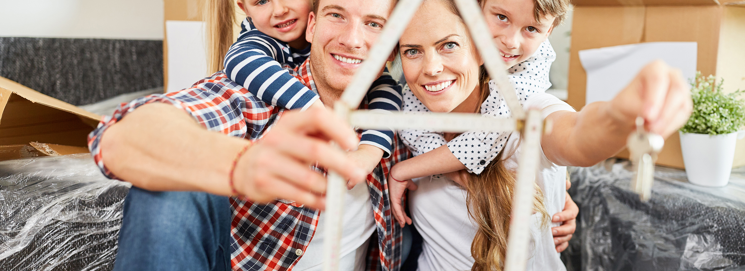 Glückliche Familie mit zwei Kindern, die zusammen ein kleines Modellhaus aus einem Zollstock formen und einen Schlüssel in der Hand halten. Symbolbild für Traumhaus für die ganze Familie.