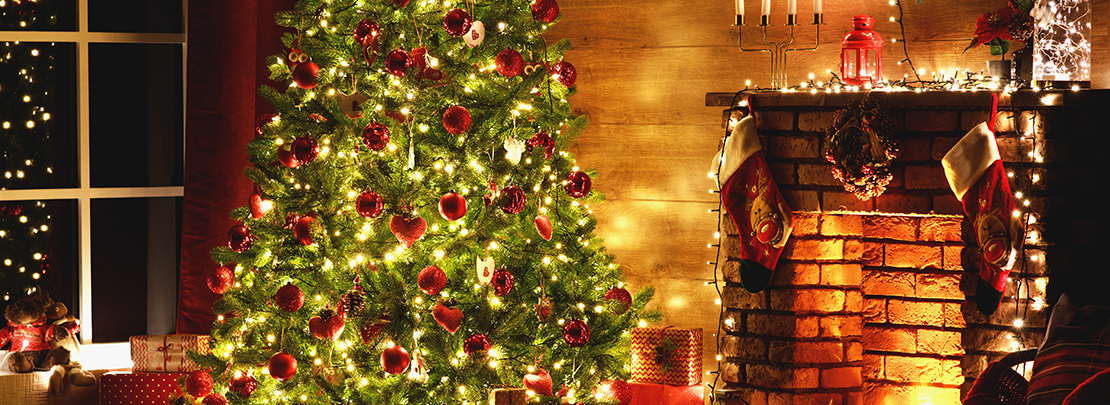 Gemütlich geschmückter Weihnachtsbaum mit Lichterketten und Geschenken vor Kamin in festlich dekoriertem Wohnzimmer.