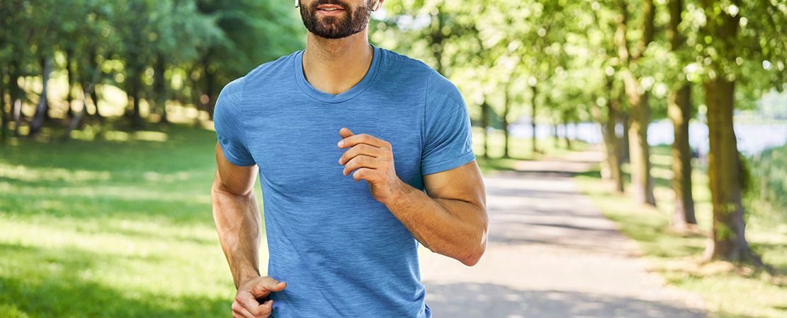 Sportlicher Mann joggt im blauen Shirt auf einem sonnigen Weg durch einen grünen Park.