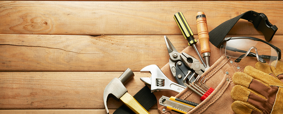 Verschiedene Handwerkzeuge wie Hammer, Schraubenzieher, Zange und Schutzbrille in einer Werkzeugtasche auf Holzhintergrund.