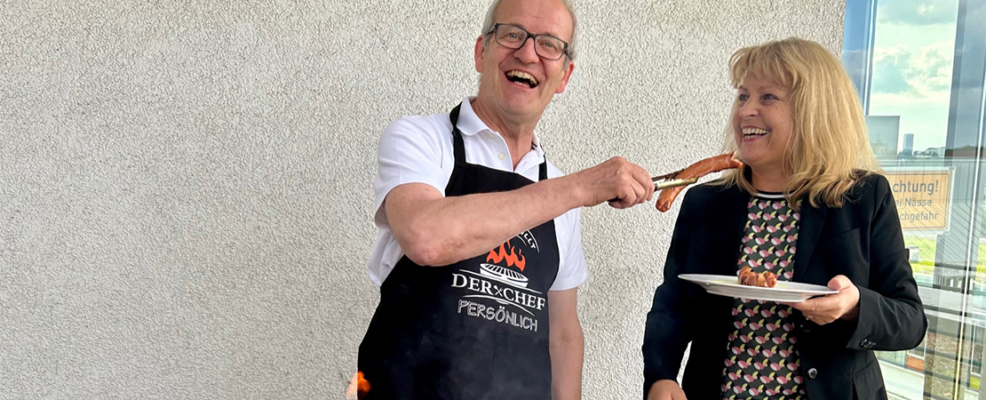 Lächelnder Mann mit Schürze „Der Chef persönlich“ reicht einer Frau mit Grillzange eine Bratwurst bei einem Afterwork Grillen.