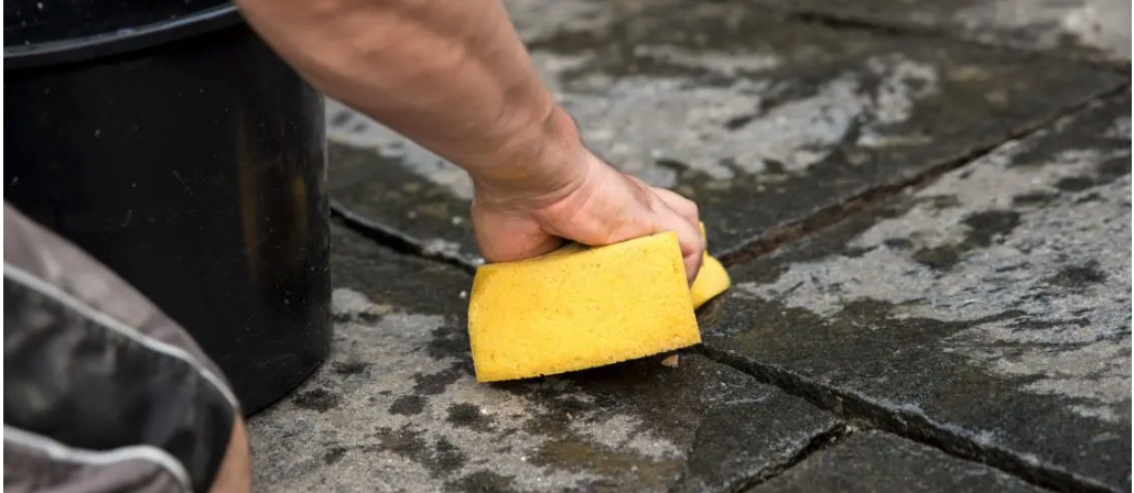 Person reinigt nasse Steinplatten mit einem gelben Schwamm, daneben steht ein schwarzer Eimer.