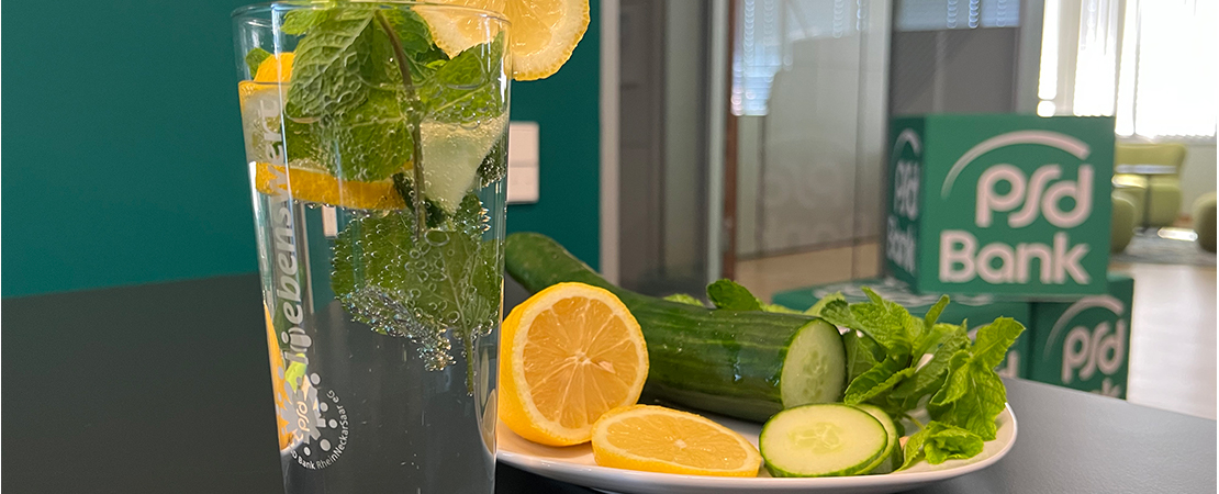 Erfrischungsgetränk mit Zitrone, Gurke und Minze im Glas, daneben frische Zutaten auf Teller vor PSD Bank Branding. Symbolbild für Tipps gegen Hitze.