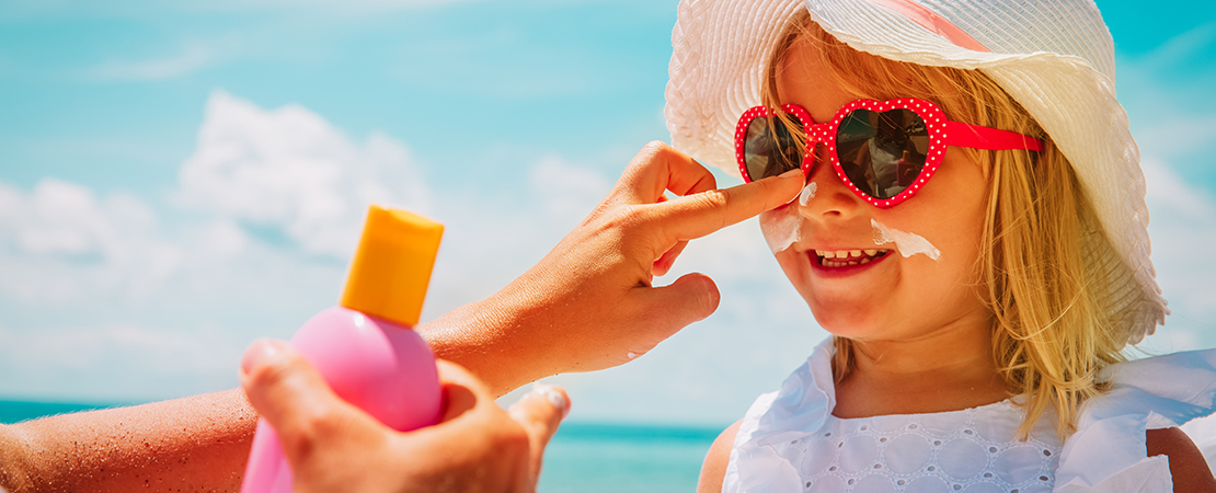 Lächelndes Mädchen mit Sonnenhut und roter Sonnenbrille bekommt am Strand Sonnencreme auf die Nase aufgetragen. Symbolbild für Sonnenschutz.