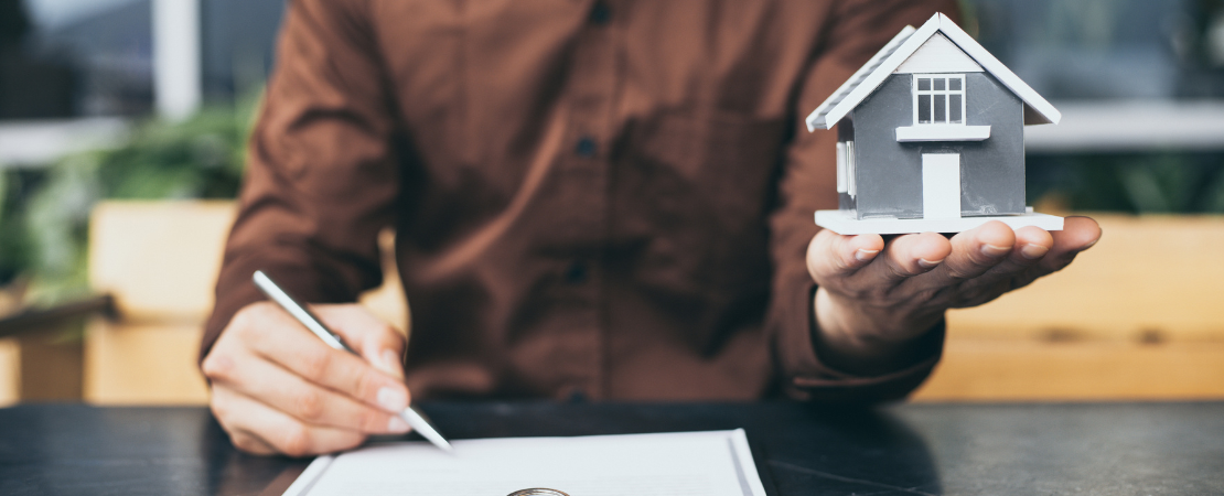 Person hält ein Miniaturmodell eines Hauses in der Hand, während er mit der anderen Hand einen Vertrag unterschreibt. Auf dem Tisch liegt ein Dokument mit einem Stift. Symbolbild für Immobilienkauf, Hausfinanzierung und Vertragsunterzeichnung beim Hauskauf.