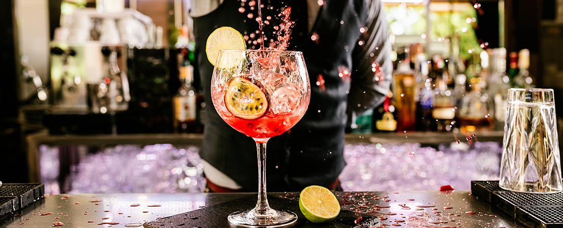 Spritziger Sommerdrink, perfekt serviert in einem bauchigen Glas mit Eiswürfeln, Limetten- und Passionsfruchtscheiben. Im Hintergrund steht ein Barkeeper vor einer stilvollen Bar, während der Drink gerade kunstvoll eingeschenkt wird – ein Moment voller Frische und Sommerlaune.