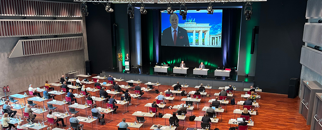 Konferenzsaal mit zahlreichen Teilnehmern an Einzeltischen, Bühne mit Rednern und großem Bildschirm mit Livestream eines Sprechers vor dem Brandenburger Tor.