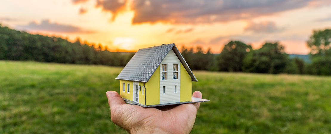 Eine Hand hält ein Modellhaus vor weitem Grünland und Sonnenuntergang deutet auf Grundstückskauf hin. Symbolbild für: Bauerwartungsland