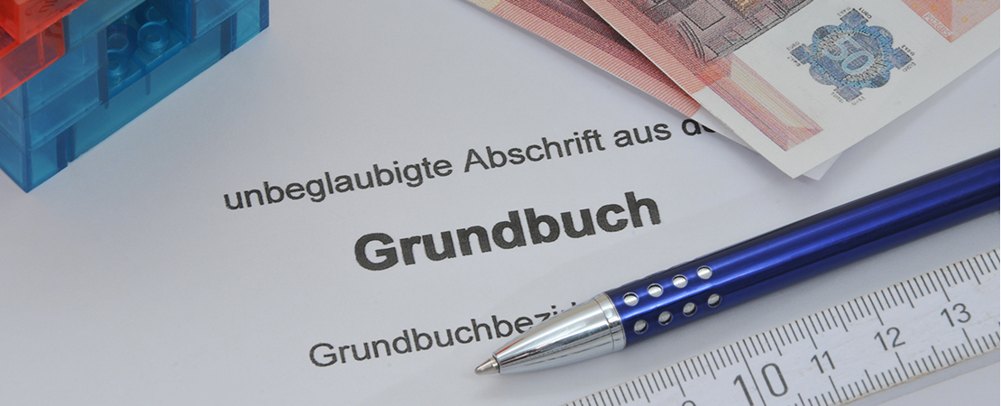 Ein Auszug aus dem Grundbuch, daneben Geldscheine, Stift und Lineal – deutet auf wichtige Informationen rund um Immobilienbesitz hin. Symbolbild für: Was steht im Grundbuch.