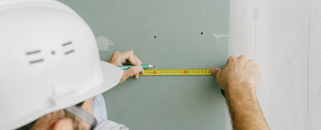 Baumängel und Mängelrügen: Ein Bauarbeiter mit Schutzhelm misst mit einem Maßband eine Wand, um Unregelmäßigkeiten oder Baufehler festzustellen. Kleine Bohrlöcher und sichtbare Unebenheiten deuten auf mögliche Baumängel hin, die überprüft oder reklamiert werden müssen.