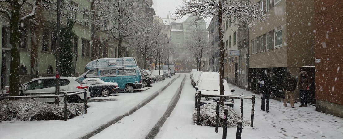 Winterliche Straßenszene in einer Stadt, bedeckt mit frischem Schnee. Autos und ein blauer Campervan sind geparkt, während Schneeflocken vom Himmel fallen. Fußgänger gehen entlang der verschneiten Gehwege, während kahle Bäume die Straße säumen. Symbolbild für: Winter, Schnee und Streusalz