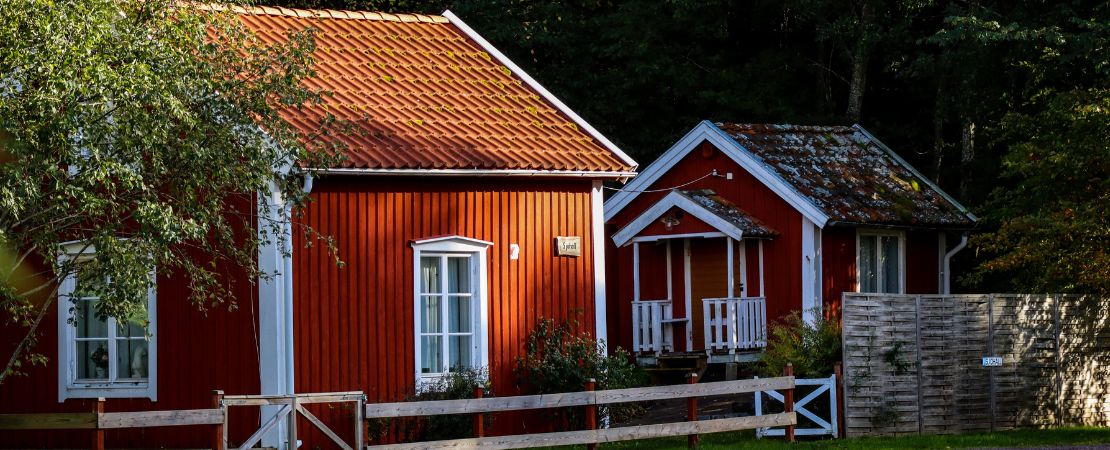 Zwei rot gestrichene Tiny Houses mit weißen Fenstern und Satteldächern in einer naturnahen Umgebung. Die kleinen Holzhäuser sind von Bäumen umgeben und durch einen Holzzaun abgegrenzt. Symbolbild für minimalistisches Wohnen, nachhaltige Architektur und Tiny-House-Bewegung.
