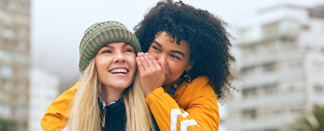 Zwei junge Frauen in warmer Kleidung lachen gemeinsam, während eine der Frauen der anderen etwas ins Ohr flüstert. Im Hintergrund sind moderne Wohngebäude zu sehen. Symbolbild für Weiterempfehlung und Prämienprogramme.