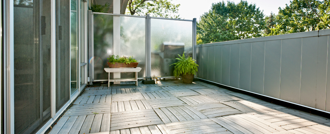 Moderne Dachterrasse mit grauem Holzboden, Sichtschutz aus Milchglas und minimalistischer Begrünung. Ein kleiner Tisch mit Pflanzkübeln und eine Topfpflanze schaffen eine gemütliche Atmosphäre. Symbolbild für urbanes Wohnen, Outdoor-Living und moderne Balkone.