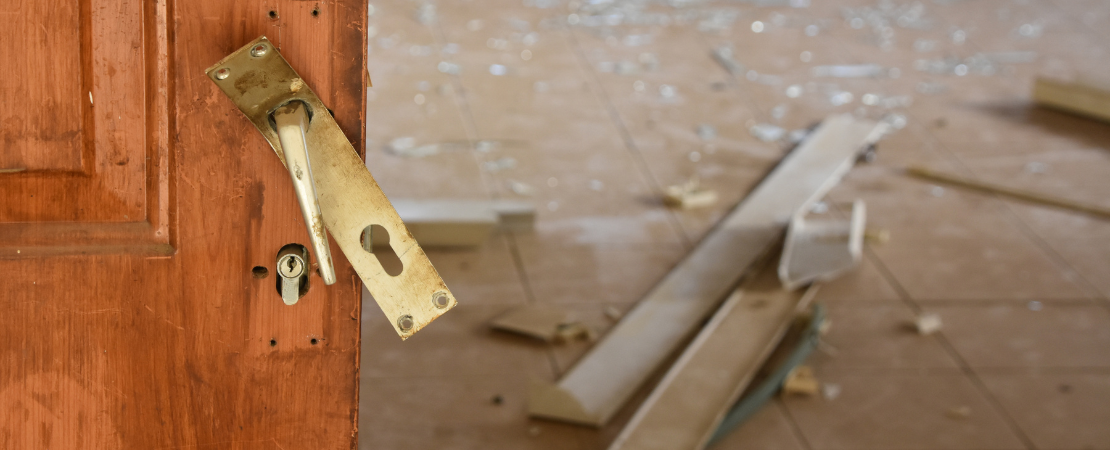 Aufgebrochene Holztür mit beschädigtem Schloss, im Hintergrund ein verwüsteter Raum mit zerbrochenem Glas und umgestürzten Möbelteilen. Symbolbild für Einbruch und Sicherheitsrisiken im Eigenheim.