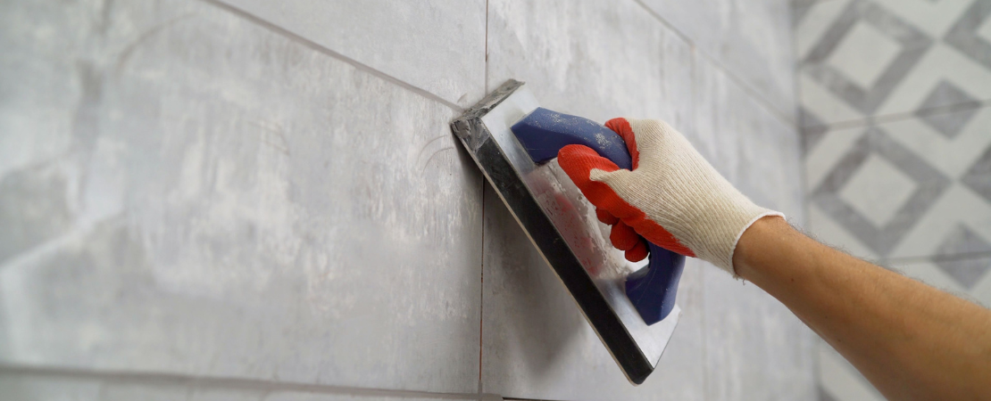 Handwerker trägt mit einem Fugbrett Fugenmasse auf eine geflieste Wand auf. Die Person trägt eine Arbeitshandschuh und verteilt das Material präzise zwischen den grauen Fliesen. Symbolbild für Fliesenverlegung, Fugen-Renovierung und Badezimmermodernisierung.
