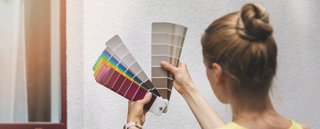 Frau hält zwei Farbfächer mit verschiedenen Farbtönen vor eine weiße Hauswand, um die passende Farbe für einen Anstrich auszuwählen. Sonnenlicht fällt auf die Oberfläche. Symbolbild für Fassadengestaltung, Farbauswahl und Renovierungsplanung.