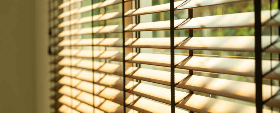 Nahaufnahme von halb geöffneten Holzjalousien an einem sonnigen Fenster. Sonnenstrahlen fallen durch die Lamellen und werfen warme Licht- und Schattenspiele auf die Fensterscheibe. Symbolbild für Sonnenschutz und natürliche Lichtregulierung im Wohnraum.