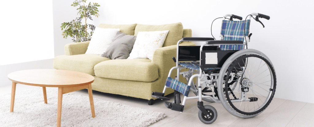 Rollstuhl mit blau-kariertem Sitzbezug steht in einem barrierefreien Wohnzimmer neben einem grünen Sofa mit dekorativen Kissen. Ein runder Holztisch und ein heller Teppich ergänzen die moderne, freundliche Einrichtung. Symbolbild für Inklusion, barrierefreies Wohnen und häusliche Pflege.