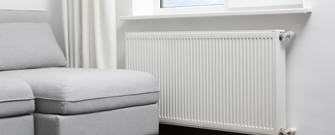 Moderner Niedrigtemperatur-Heizkörper unter einem Fenster in einem hell eingerichteten Wohnzimmer. Daneben steht ein graues Sofa mit Stoffbezug, eine weiße Gardine hängt im Hintergrund. Symbolbild für energieeffizientes Heizen, moderne Heiztechnik und Wohnkomfort.