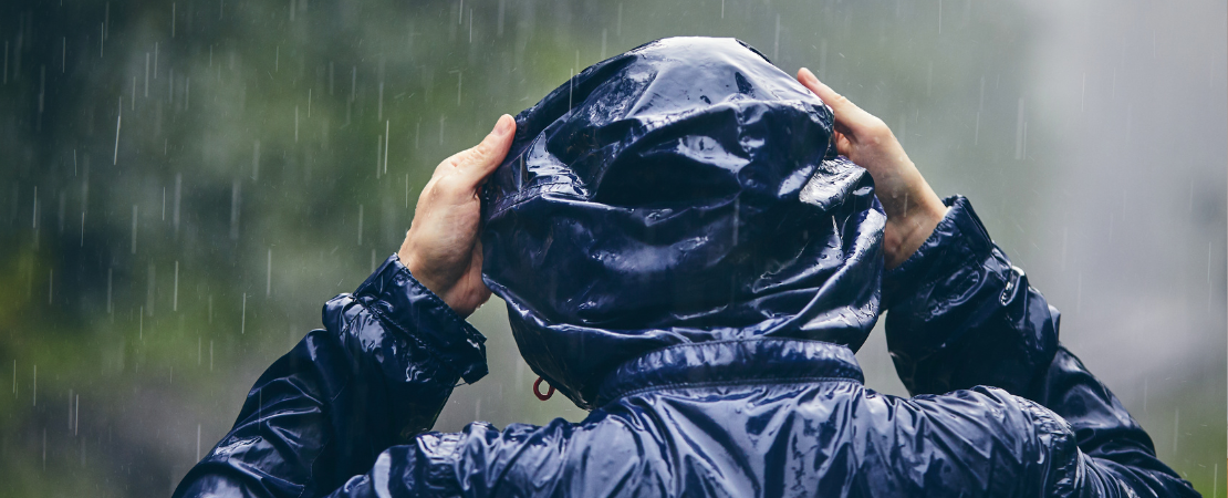 Person in einer dunkelblauen, wasserabweisenden Regenjacke zieht die Kapuze über den Kopf, während starker Regen fällt. Die nasse Kleidung glänzt unter den Regentropfen, der Hintergrund ist unscharf mit Bäumen und Natur. Symbolbild für schlechtes Wetter, Unwetter und Wetterschutz.