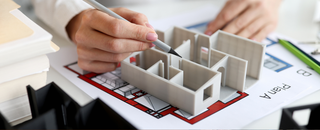 Bauplaner arbeitet an einem Aufteilungsplan einer Wohnung. Eine Hand hält einen Bleistift und zeigt auf eine Wand des Miniatur-Modells. Im Hintergrund liegen Akten, Planungsdokumente und ein Klemmbrett.