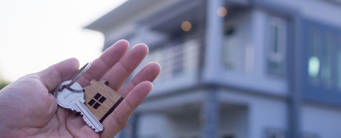 Hand hält einen Schlüsselbund mit einem Haus-Schlüsselanhänger vor einem modernen Wohnhaus. Unscharfer Hintergrund mit beleuchteten Fenstern symbolisiert Hauskauf, Immobilienbesitz oder Mietbeginn. Konzeptbild für Eigenheim, Immobilienmarkt und Neubau.