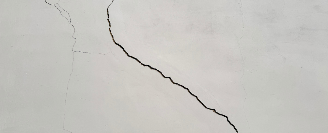 Großer Riss in einer weißen Betonwand. Die unregelmäßige Struktur des Risses zieht sich diagonal über die Oberfläche und symbolisiert Bauschäden, Materialermüdung oder strukturelle Probleme.