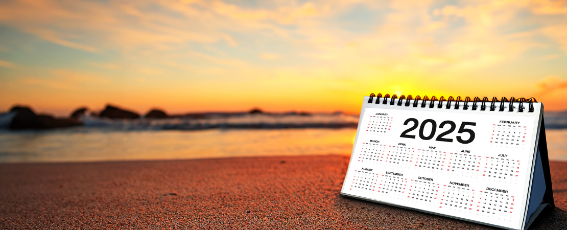 Tischkalender mit der Jahreszahl 2025 auf einem Sandstrand bei Sonnenuntergang. Im Hintergrund trifft das Meer auf die Küste, während der Himmel in warmen Farben leuchtet. Symbolbild für die Urlaubsplanung 2025.