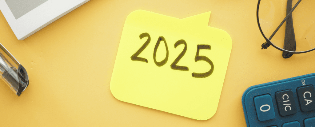 Gelber Notizzettel in Form einer Sprechblase mit handgeschriebener Jahreszahl '2025' auf einem gelben Schreibtisch. Daneben befinden sich eine Brille, ein Taschenrechner, ein Kugelschreiber und ein Notizbuch. Symbolbild für Zukunft, Planung und Geschäftsjahr 2025.