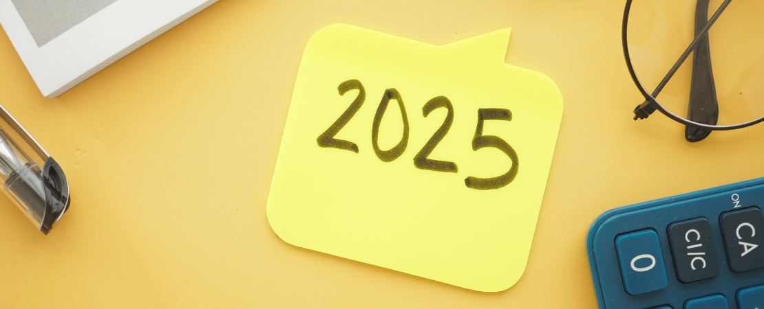 Gelber Notizzettel in Form einer Sprechblase mit handgeschriebener Jahreszahl '2025' auf einem gelben Schreibtisch. Daneben befinden sich eine Brille, ein Taschenrechner, ein Kugelschreiber und ein Notizbuch. Symbolbild für Zukunft, Planung und Geschäftsjahr 2025.