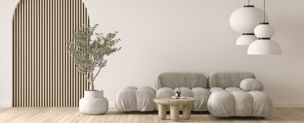Modernes Wohnzimmer mit minimalistischem Design: Eine graue, organisch geformte Couch, ein runder Holztisch und eine große Zimmerpflanze in einem Beton-Topf stehen auf hellem Holzboden. Eine dekorative Holzlamellenwand mit rundem Durchgang und drei stilvolle Papierlampen verleihen dem Raum eine warme, skandinavische Ästhetik.