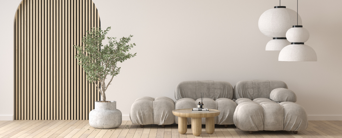 Modernes Wohnzimmer mit minimalistischem Design: Eine graue, organisch geformte Couch, ein runder Holztisch und eine große Zimmerpflanze in einem Beton-Topf stehen auf hellem Holzboden. Eine dekorative Holzlamellenwand mit rundem Durchgang und drei stilvolle Papierlampen verleihen dem Raum eine warme, skandinavische Ästhetik.